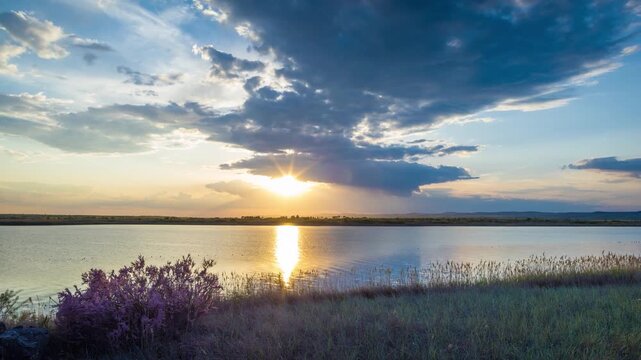 Clouds run across the sky with a sunset light over a lake with herbs on a shore