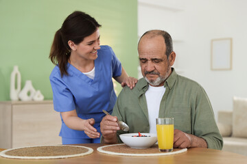Caregiver giving breakfast to elderly man at wooden table indoors