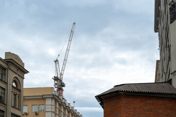 Fototapeta premium Crane Towering Over City Rooftops, Cloudy Sky, Brick Facades, Historic Buildings Undergoing Renovation, Crane Boom And Hook Poised, Materials Hoisted