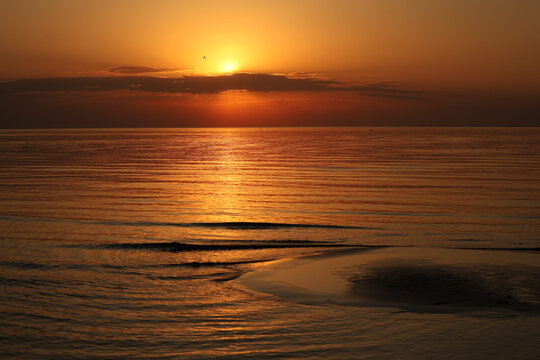 Beautiful golden hour seascape with a bright setting sun piercing through the cloudline, casting a luminous path over the rippling water and shore.