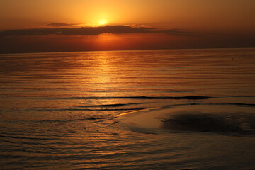Fototapeta premium Golden setting sun peeking from under a dark cloud bank, creating a bright shimmering reflection on the calm sea and a shallow sandbank.