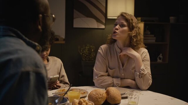 Candid over shoulder shot of parents fighting while sitting at dinner table at home with focus on upset adult woman talking and leaving room, scene lit by low evening lighting