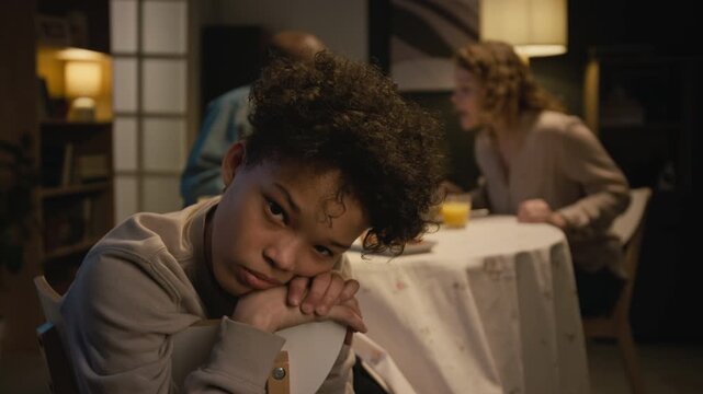 Close up shot of sad Black teenage boy looking at camera with parents fighting at dinner table in blurred background, dysfunctional family dynamics, copy space