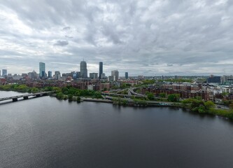 Obraz premium Aerial drone view of the Boston skyline under a cloudy sky, featuring the Charles River, Harvard Bridge, and Back Bay brownstones leading toward the Prudential and John Hancock towers.