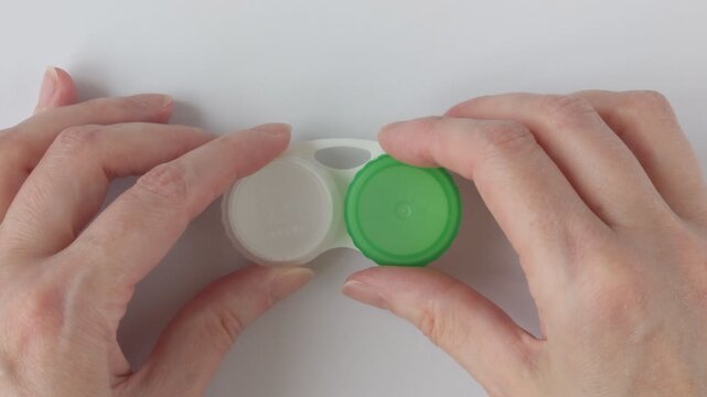 Woman's hands carefully unscrew the green lid of an empty contact lens case. Empty white plastic case with compartments for storing contact lenses. Concept for eye care and hygienic lens storage