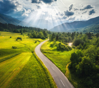 Aerial view of road in green meadows at sunset in summer. Top view from drone of rural road, mountains, forest. Beautiful landscape with roadway, sun rays, trees, hills, green grass, clouds. Slovenia