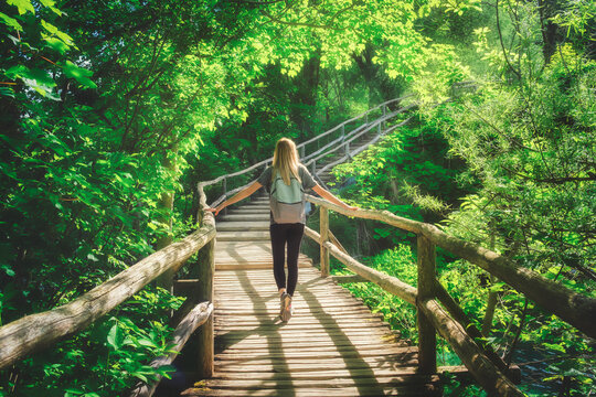 Young woman walking on wooden boardwalk over turquoise lakes in Plitvice Lakes National Park, Croatia. Female tourist enjoying green forest during summer trip. Traveler on wooden trail and green trees