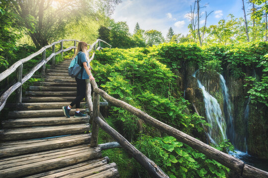 Young woman with backpack with backpack standing on wooden stairs near waterfalls, lush green trees in Plitvice Lakes national park, Croatia at sunset in summer. Female tourist. Girl in forest. Hiking