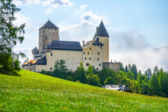 Mauterndorf Castle stands majestically among lush greenery in the Austrian Alps, showcasing medieval architecture against a backdrop of clear blue skies and distant mountains.