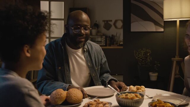 Medium shot of Black man as happy father enjoying dinner with multiracial family and saying grace while sitting at table together, cozy evening scene lit by low lighting, copy space