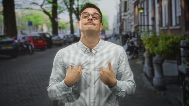 Man pointing thumbs to chest on a cobblestone street lined with parked cars and bicycles, wearing striped long sleeve shirt and glasses, slight smile and hesitant expression; pleading apology.