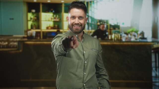 Young hispanic man with beard points finger at camera inside a bar building, smiling and leaning forward; confidence welcome.