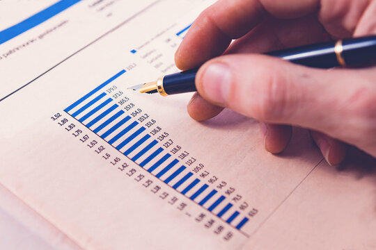 Mano sosteniendo una pluma estilogr&aacute;fica y se&ntilde;alando un informe financiero con gr&aacute;fico de barras. Primer plano de un an&aacute;lisis empresarial. Un profesional analiza un informe financiero para decisiones 