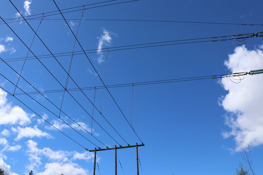 Detailed view of a high-voltage power line, captured against a clear blue sky with bright sunlight and scattered cumulus clouds. Contemporary Swedish energy infrastructure presented in sharp daylight 