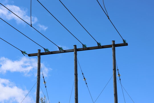 Detailed view of a high-voltage power line, captured against a clear blue sky with bright sunlight and scattered cumulus clouds. Contemporary Swedish energy infrastructure presented in sharp daylight 