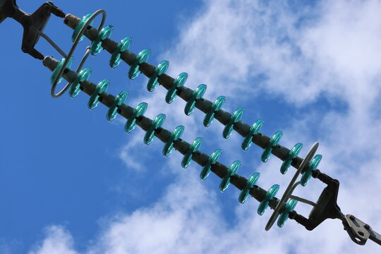 Detailed view of a high-voltage power line, captured against a clear blue sky with bright sunlight and scattered cumulus clouds. Contemporary Swedish energy infrastructure presented in sharp daylight 