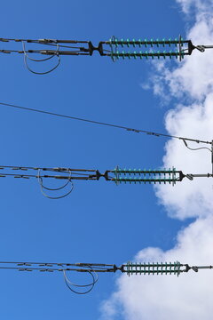 Detailed view of a high-voltage power line, captured against a clear blue sky with bright sunlight and scattered cumulus clouds. Contemporary Swedish energy infrastructure presented in sharp daylight 