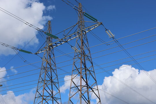 Detailed view of a high-voltage power line, captured against a clear blue sky with bright sunlight and scattered cumulus clouds. Contemporary Swedish energy infrastructure presented in sharp daylight 