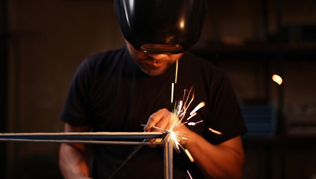 A young male welder wearing a black t-shirt and protective helmet bends over a steel tube frame, striking a bright spark burst with a welding rod in a warm dimly lit fabrication workshop.