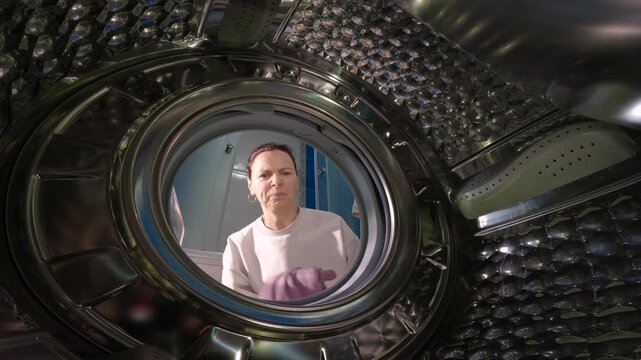 Woman loading dirty clothes into washing machine drum. Unique perspective from inside the washing machine drum showing a woman opening the door, putting dirty clothes inside