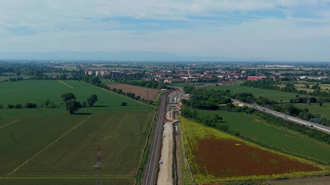 Railway Construction in Rural Landscape: An aerial view captures the ongoing construction of a new railway line cutting through a serene rural landscape, bordered by green fields and a distant town.