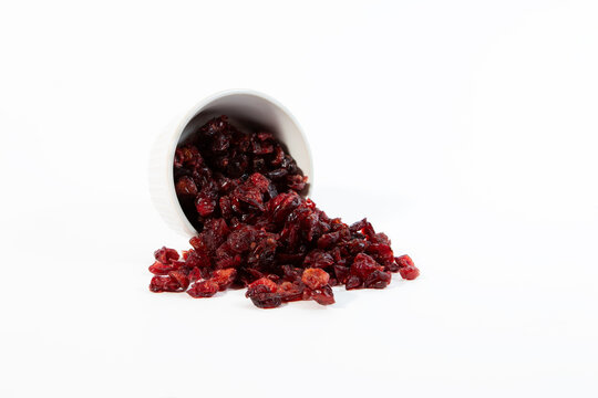 Dried cranberries spilled out from a tipped white ceramic cup onto a clean white background