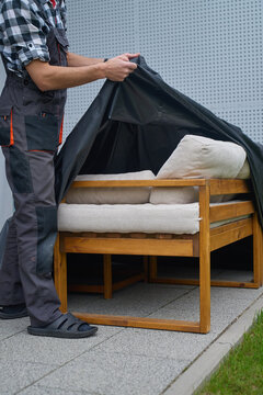 Man removes protective cover from outdoor sofa with cushions on patio. Concept of garden furniture care