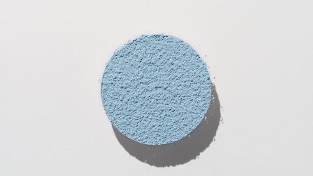 Overhead frame of pale blue powder sifting into a circle casts a fluctuating shadow on a pale grey background