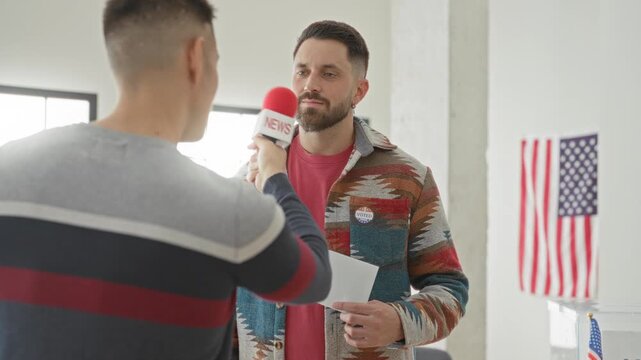 Man holds news microphone while another man displays an i voted sticker and ballot inside a polling station with us flag visible; pride civic duty.