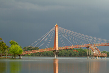 Suspension bridge over calm river under dark cloudy sky, landscape with reflection, trees, and...