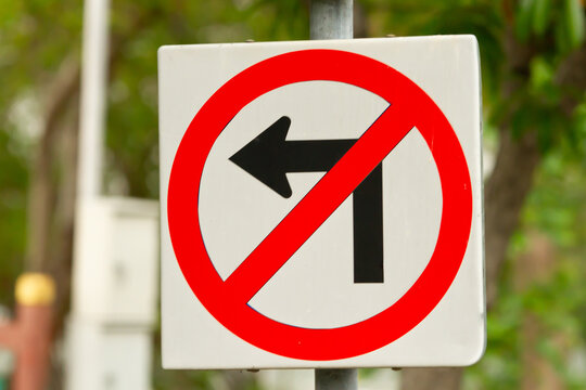 No left turn sign on white traffic sign with red circle and diagonal slash, outdoor road safety symbol with blurred green background
