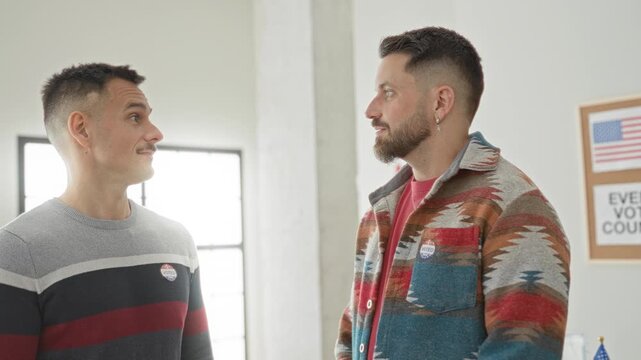 Two men talking, one clasping hands and both wearing 'i voted' stickers in a polling building; civic duty pride.