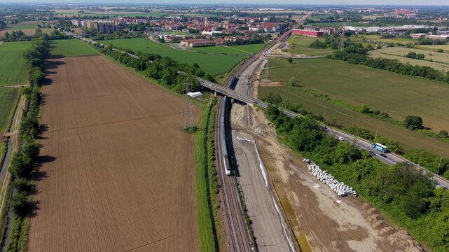Aerial View of Infrastructure: An aerial perspective captures a modern highway intersection with trains passing on parallel tracks, surrounded by agricultural fields and distant buildings.