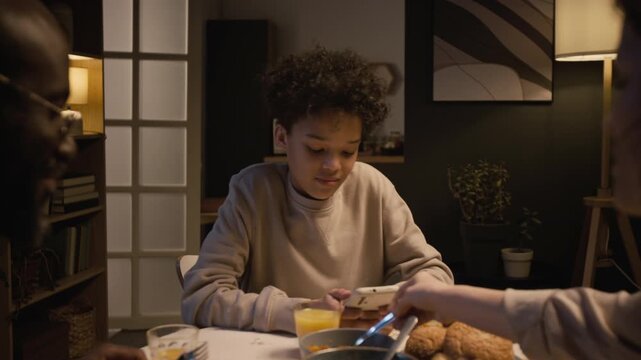Medium shot of distracted Black teenage boy using smartphone at dinner table with family and smiling reading messages, gadget addiction in children, copy space