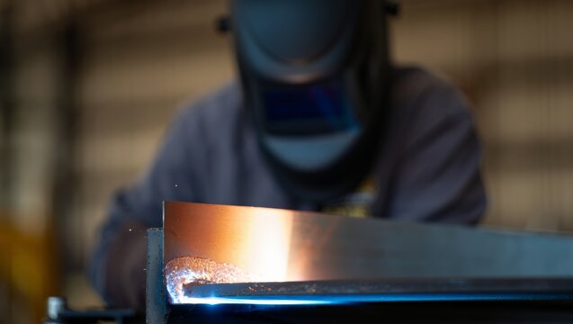 Shallow focus shot of a freshly formed weld bead glowing orange on a steel girder edge, with the helmeted welder in gray worksuit softly blurred in the warm background of an industrial workshop.