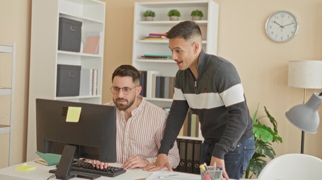 Two men at a computer, seated worker raising hand for fistbump while standing colleague leans over the desk in building; teamwork camaraderie.