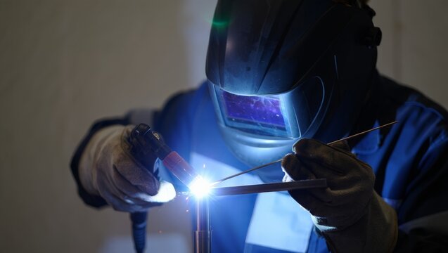 Close portrait of a welder in blue work jacket and large auto-darkening helmet with glowing purple visor reflection, TIG welding with a filler rod with bright white arc in a dim workshop.