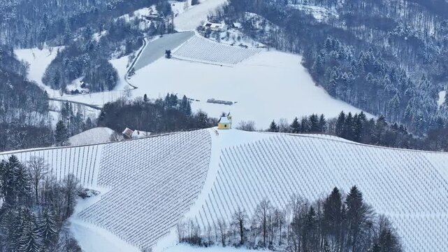Aerial view of Dreisiebner Kapelle on a snow-covered hill surrounded by vineyards and evergreen forests in Gamlitz, Styria, Austria.