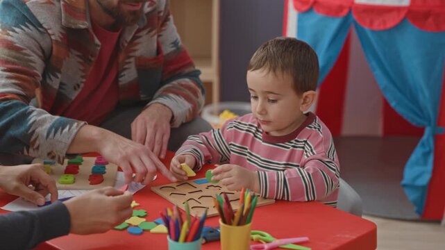 Boy fitting colored shapes into a wooden puzzle on a red table, hands guiding him, crayons and scissors visible in a playroom building; playful learning joy.