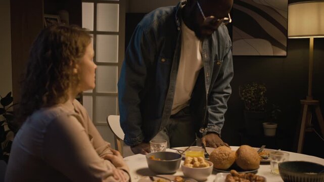Side view shot of multiracial couple fighting at dinner table with Black man storming off pushing furniture and leaving upset wife alone, scene lit by low evening light, copy space