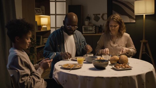 Gadget addiction in children, strict father talking to teenage boy using smartphone at dinner table with family, slamming hand on table and boy storming off, copy space