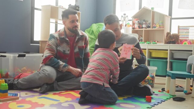 Boy kneeling while two men on floor show flashcards and point at pictures in a preschool classroom building; play learning joy.