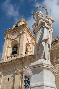Low angle vertical view of 
statue of Virgin Mary with halo. 
Ancient catholic church bell tower 
with clock on background in 
Cospicua Bormla, Malta.
