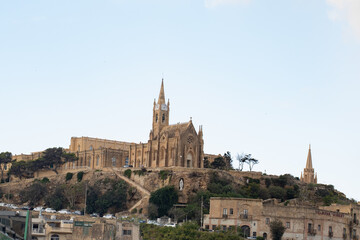 Fototapeta premium Horizontal scenic view of ancient neo-gothic church on rocky hill. Majestic old limestone cathedral architecture background in Gozo island, Malta. 