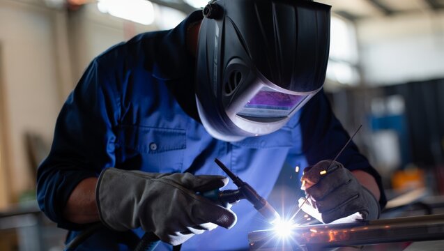 Close-up of a professional TIG welder in blue work shirt and large black auto-darkening helmet with gray TIG gloves welding a precise metal joint with a bright white arc in a workshop.