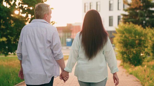 Mature, happy couple strolls hand in hand on date in city park. Man woman walking together on date nature park. Loving husband wife relaxing outdoors in summer, enjoying time together. Love tenderness