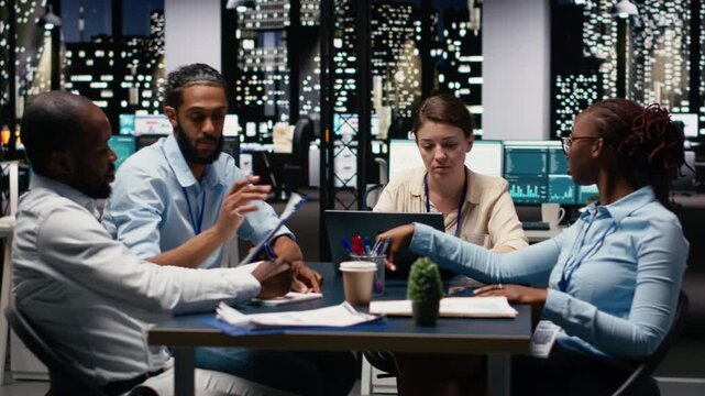 Diverse team of employees in office looking over paperwork documents late at night. Coworkers at work cooperating, analyzing company budget reports documentation files before deadline