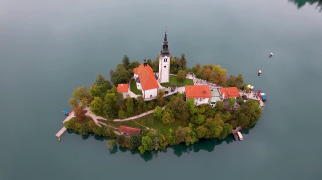 Aerial view of Lake Bled island featuring the Pilgrimage Church of the Assumption of Mary with people walking on the paths surrounded by calm water in Bled, Radovljica, Slovenia.