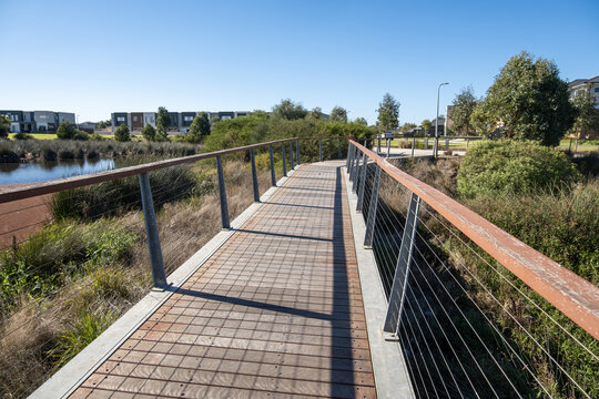 A pedestrian bridge spans a suburban wetland in Point Cook, Melbourne Australia. The bridge path leads toward a landscaped parkland trail with native vegetation, a retention pond and townhouses.