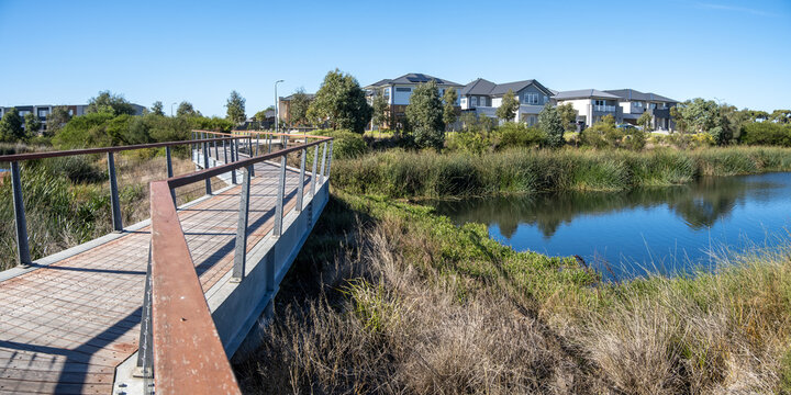 Wooden pedestrian boardwalk over suburban wetland and retention pond with modern two-story family houses in the background, urban planning and green infrastructure in Point Cook, Melbourne, Australia